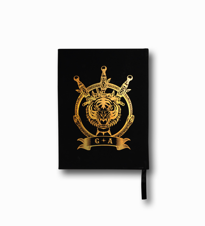 THE ALCHEMIST JOURNAL by GARDEN ALCHEMY is a black A5 vegan leather journal featuring a gold crowned tiger head with swords and a circle, plus G A on a ribbon. It includes a black ribbon bookmark.