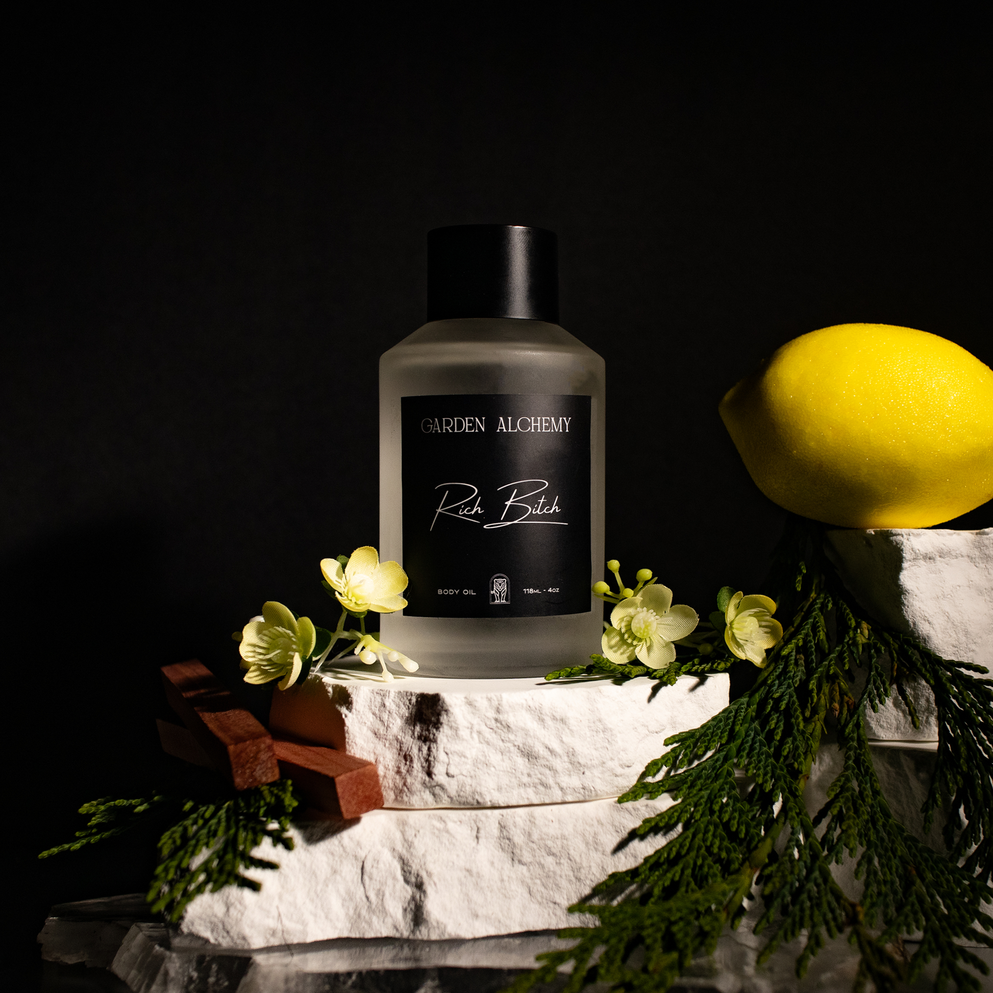 A bottle of GARDEN ALCHEMY RICH BITCH BODY OIL rests on white stone, with green leaves, yellow flowers, lemon, and wood fragments arranged nearby against a dark background.