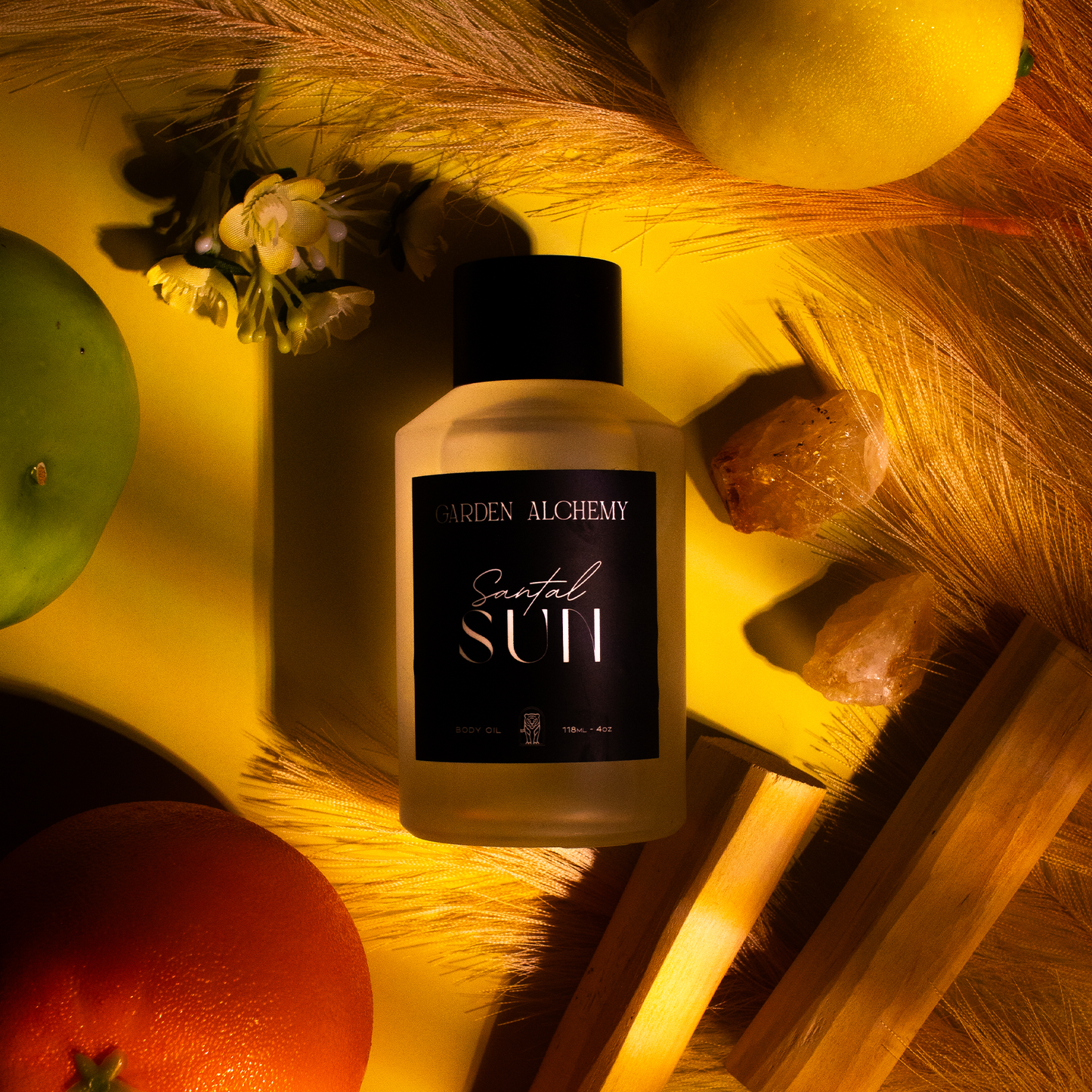 A bottle of GARDEN ALCHEMY SANTAL SUN BODY OIL is surrounded by fruit, crystals, white flowers, wooden sticks, and dried grasses, all bathed in warm yellow light.