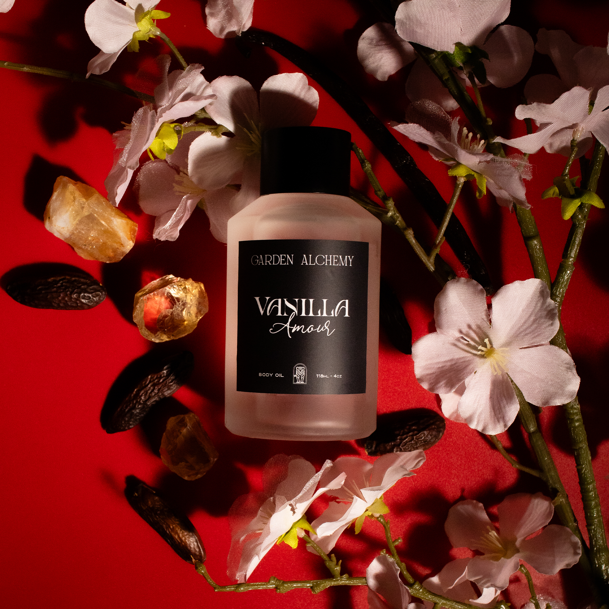 A bottle of GARDEN ALCHEMY VANILLA AMOUR w/pheromones, with an oriental gourmand scent, sits amid pink cherry blossoms, resin pieces, and dark vanilla beans on a deep red background.